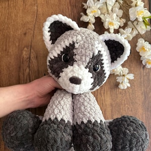 May include: A handmade crochet raccoon plush toy. The stuffed animal features a gray and white body with black accents around the eyes, ears, and feet. The plush toy is soft and cuddly, perfect for children or as a decorative item.