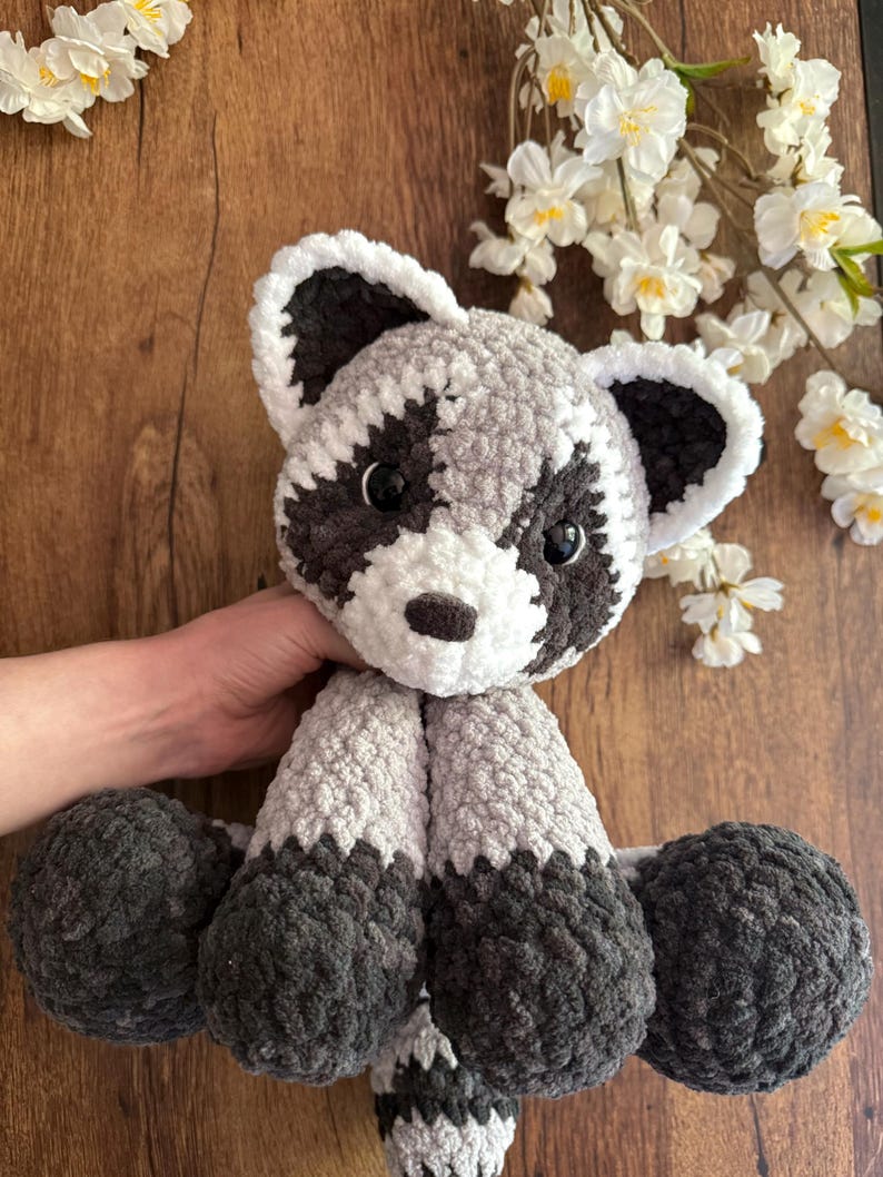 May include: A handmade crochet raccoon plush toy. The stuffed animal features a gray and white body with black accents around the eyes, ears, and feet. The plush toy is soft and cuddly, perfect for children or as a decorative item.