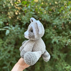May include: A handmade gray and white crochet teddy bear. The plush toy has a soft, textured appearance with a light gray body, white accents on the ears and face, and dark gray paws. The bear is held against a backdrop of green foliage.