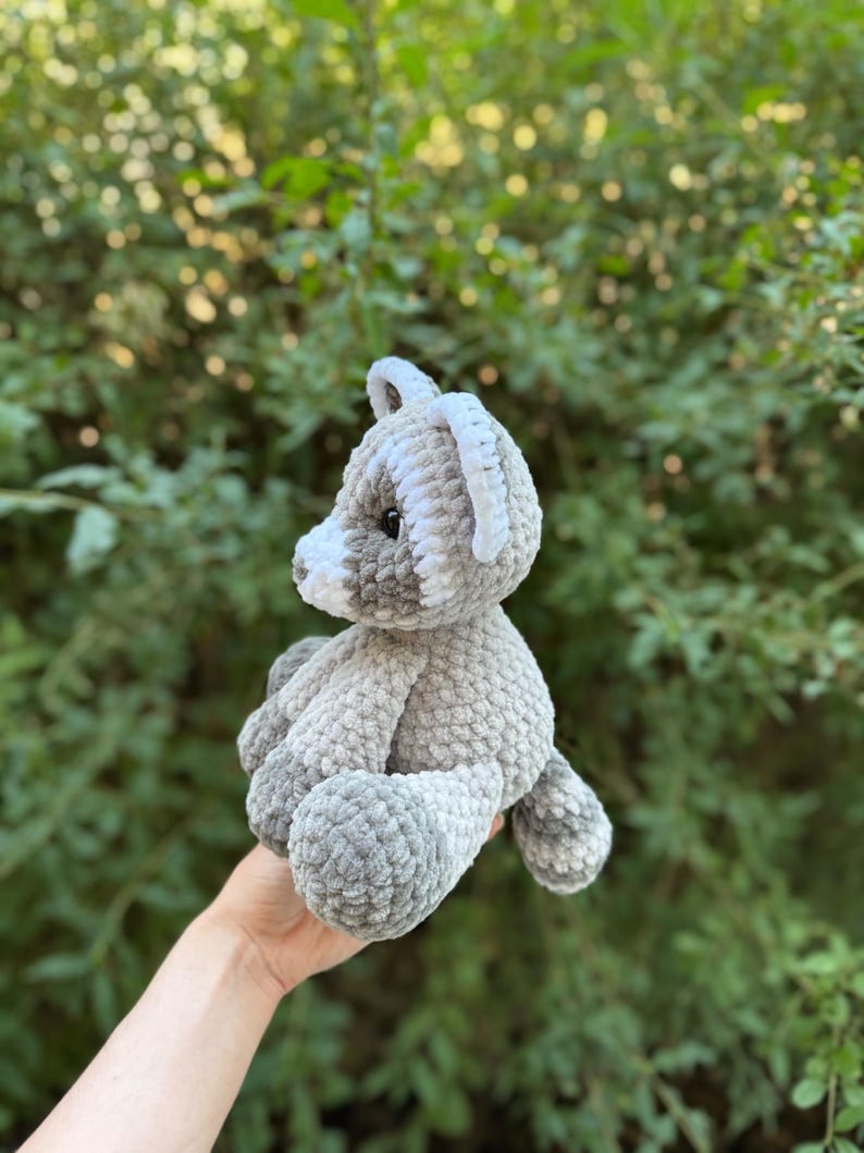 May include: A handmade gray and white crochet teddy bear. The plush toy has a soft, textured appearance with a light gray body, white accents on the ears and face, and dark gray paws. The bear is held against a backdrop of green foliage.