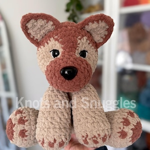 May include: A plush crochet dog toy featuring a textured tan and brown yarn body, large pointed ears, black plastic eyes, and a black nose. The text Knots and Snuggles is overlaid across the center of the stuffed animal in white lettering.