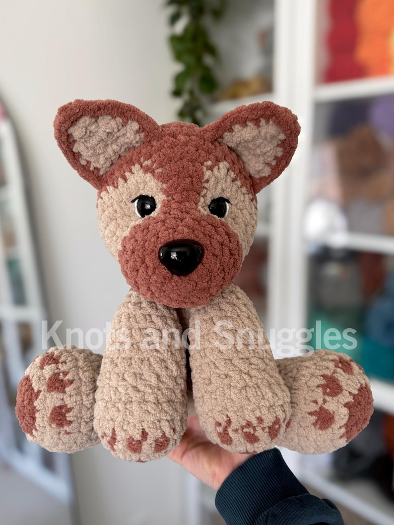 May include: A plush crochet dog toy featuring a textured tan and brown yarn body, large pointed ears, black plastic eyes, and a black nose. The text Knots and Snuggles is overlaid across the center of the stuffed animal in white lettering.