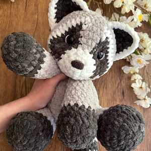 May include: A handmade crochet raccoon plush toy. The stuffed animal features a gray and white body with dark gray accents on the paws, tail, and around the eyes. The raccoon has black eyes and a white muzzle. The plush is soft and cuddly.