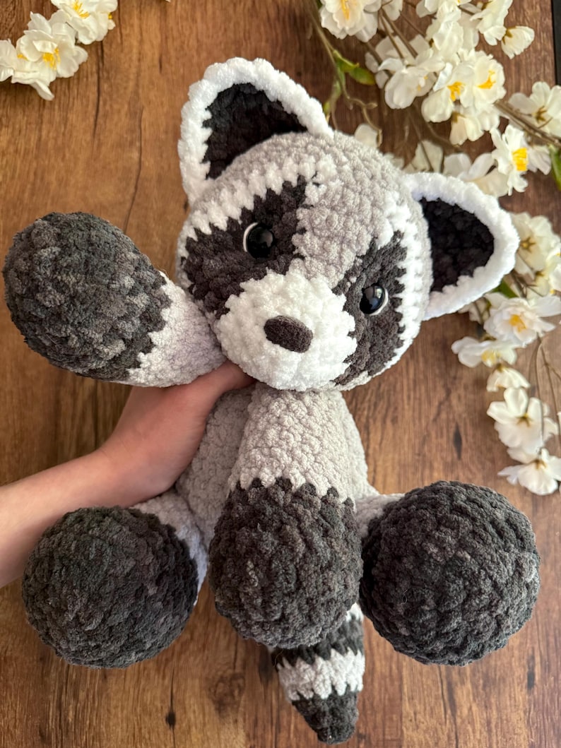 May include: A handmade crochet raccoon plush toy. The stuffed animal features a gray and white body with dark gray accents on the paws, tail, and around the eyes. The raccoon has black eyes and a white muzzle. The plush is soft and cuddly.