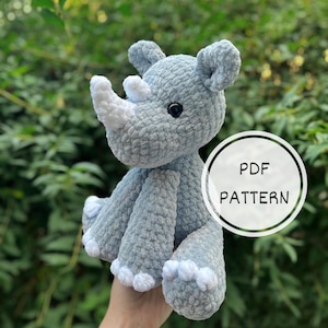 Pode incluir: Um brinquedo de pelúcia de rinoceronte cinza e branco feito de crochê. O brinquedo está sentado em uma mão e tem um chifre branco. O texto "PDF PATTERN" está em um círculo branco atrás do brinquedo.