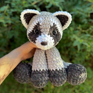 May include: A handmade crochet raccoon plush toy. The raccoon has a gray and white body with black markings around the eyes and on the tail. The plush is soft and cuddly, perfect for children or as a decorative item.