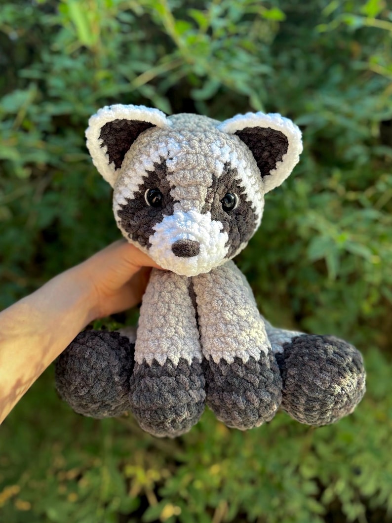 May include: A handmade crochet raccoon plush toy. The raccoon has a gray and white body with black markings around the eyes and on the tail. The plush is soft and cuddly, perfect for children or as a decorative item.