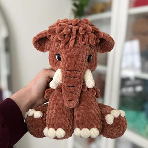 May include: A handmade, plush mammoth toy in shades of brown and cream. The stuffed animal has a shaggy head, large ears, and white tusks. The toy is soft and cuddly, perfect for children.