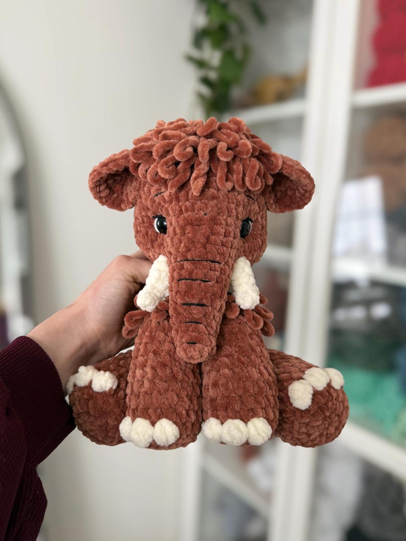 May include: A handmade, plush mammoth toy in shades of brown and cream. The stuffed animal has a shaggy head, large ears, and white tusks. The toy is soft and cuddly, perfect for children.
