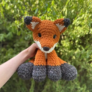 Puede incluir: Un peluche de zorro tejido a ganchillo, con detalles en naranja, blanco y gris. El zorro tiene un aspecto texturizado, con hocico blanco, ojos negros y patas grises. El fondo es follaje verde.