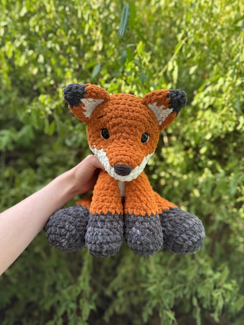Puede incluir: Un peluche de zorro tejido a ganchillo, con detalles en naranja, blanco y gris. El zorro tiene un aspecto texturizado, con hocico blanco, ojos negros y patas grises. El fondo es follaje verde.