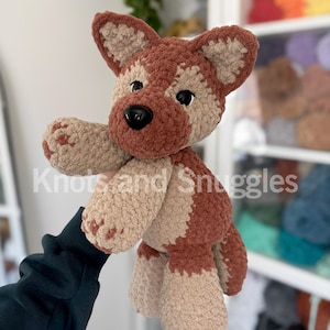 May include: A plush crochet dog toy featuring a soft, textured yarn in shades of brown and beige. The stuffed animal has large, pointed ears, a dark brown snout, and black plastic safety eyes. The text Knots and Snuggles is overlaid on the image.