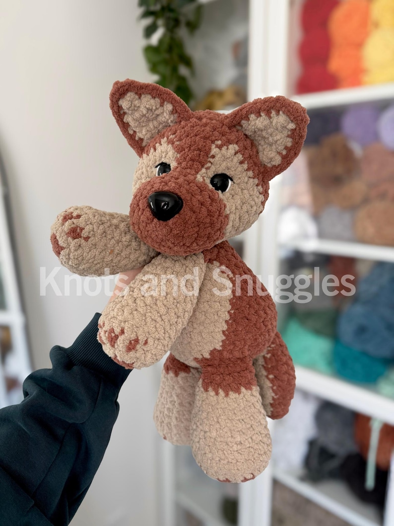May include: A plush crochet dog toy featuring a soft, textured yarn in shades of brown and beige. The stuffed animal has large, pointed ears, a dark brown snout, and black plastic safety eyes. The text Knots and Snuggles is overlaid on the image.