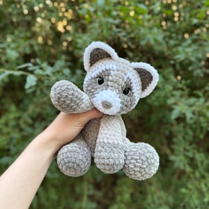May include: A handmade crochet raccoon plush toy in shades of gray and white. The stuffed animal has a white muzzle, ears, and accents, with black eyes. The plush toy is soft and cuddly, perfect for children or as a decorative item.