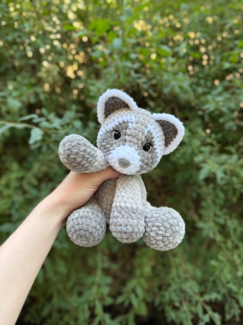 May include: A handmade crochet raccoon plush toy in shades of gray and white. The stuffed animal has a white muzzle, ears, and accents, with black eyes. The plush toy is soft and cuddly, perfect for children or as a decorative item.
