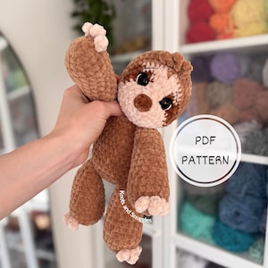 Puede incluir: Un peluche de perezoso tejido a ganchillo en marrón y beige con grandes ojos de seguridad negros y garras claras. El juguete presenta una superposición gráfica circular con el texto PDF PATTERN y el texto Knots and Snuggles en la pierna.
