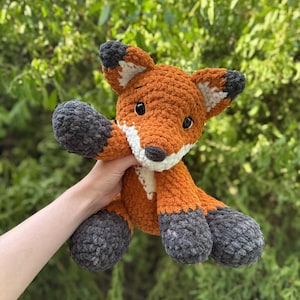 Puede incluir: Un peluche de zorro tejido a ganchillo con detalles en naranja, blanco y gris. El zorro tiene una textura esponjosa, ojos negros y est&aacute; posado con los brazos extendidos. El fondo es un follaje verde borroso.