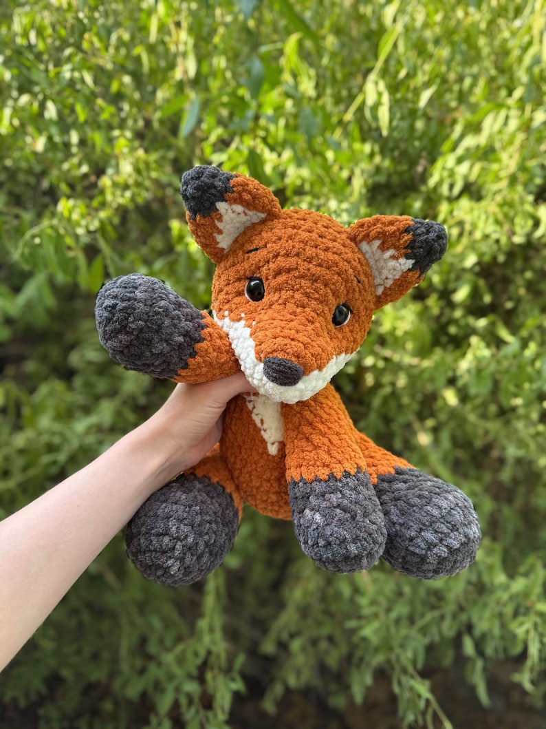Puede incluir: Un peluche de zorro tejido a ganchillo con detalles en naranja, blanco y gris. El zorro tiene una textura esponjosa, ojos negros y est&aacute; posado con los brazos extendidos. El fondo es un follaje verde borroso.