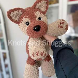 May include: A plush crochet dog toy featuring a soft, textured yarn in shades of brown and beige. The stuffed animal has large black plastic eyes, a black nose, and pointed ears. The text Knots and Snuggles is visible across the center.