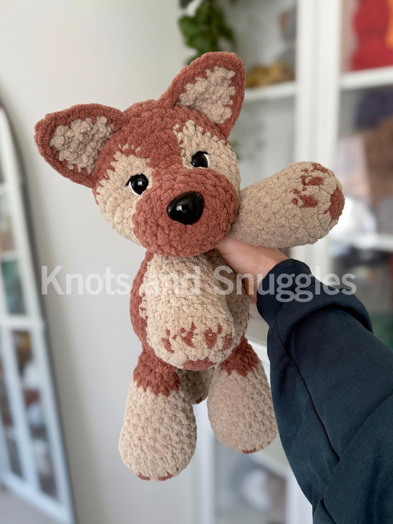 May include: A plush crochet dog toy featuring a soft, textured yarn in shades of brown and beige. The stuffed animal has large black plastic eyes, a black nose, and pointed ears. The text Knots and Snuggles is visible across the center.