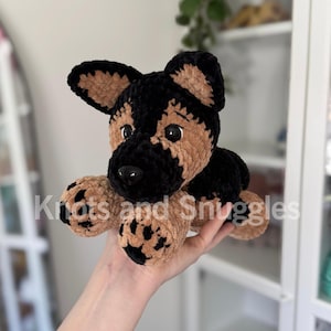 May include: A plush German Shepherd dog toy made of soft, textured yarn in black and tan colors. The toy features large black plastic eyes, a black nose, and paw pads with black detailing. The text Knots and Snuggles is overlaid on the image.