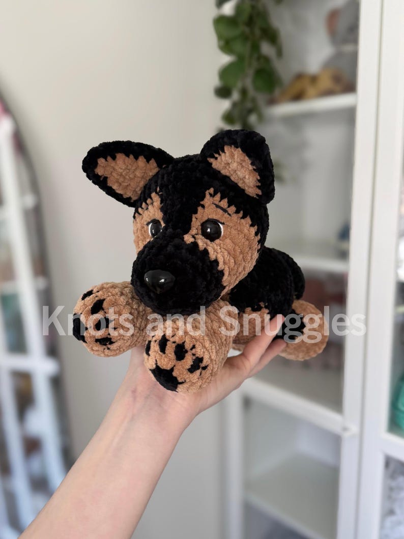 May include: A plush German Shepherd dog toy made of soft, textured yarn in black and tan colors. The toy features large black plastic eyes, a black nose, and paw pads with black detailing. The text Knots and Snuggles is overlaid on the image.