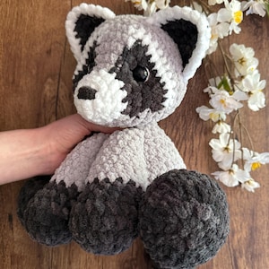 May include: A handmade crochet raccoon plush toy. The plush features a gray and white body with black accents around the eyes, ears, and feet. The raccoon has a striped tail and is made of soft, textured yarn.