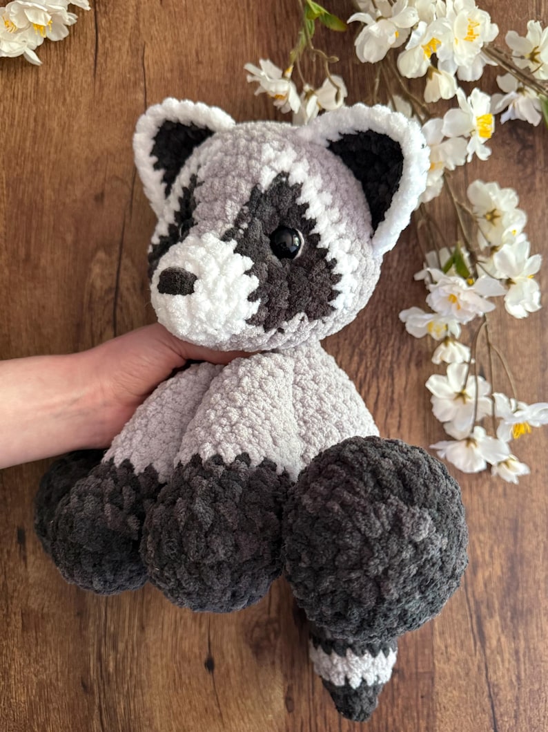 May include: A handmade crochet raccoon plush toy. The plush features a gray and white body with black accents around the eyes, ears, and feet. The raccoon has a striped tail and is made of soft, textured yarn.