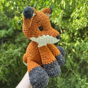 Puede incluir: Un peluche de zorro hecho a mano con ganchillo. El zorro es principalmente naranja con detalles blancos en el pecho y el hocico. Las orejas y los pies son de color gris oscuro. El zorro est&aacute; sentado.