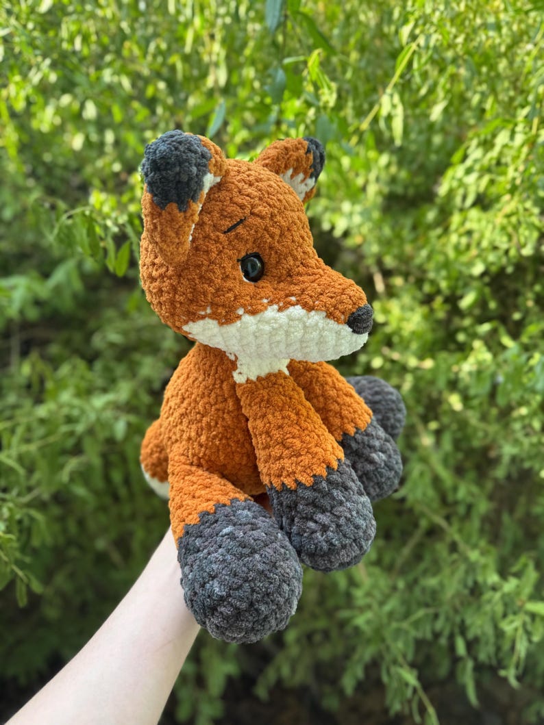 Puede incluir: Un peluche de zorro hecho a mano con ganchillo. El zorro es principalmente naranja con detalles blancos en el pecho y el hocico. Las orejas y los pies son de color gris oscuro. El zorro est&aacute; sentado.