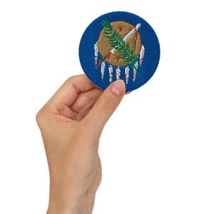 May include: A circular, blue embroidered patch displaying the Oklahoma state seal. The seal features a central shield with a Native American design, a peace pipe, and olive branches. The patch is held in a hand against a white backdrop.