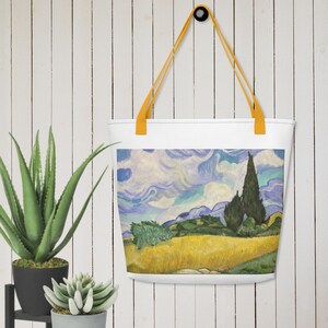 Könnte beinhalten: Eine weiße Canvas-Tragetasche mit gelben Henkeln. Die Tasche zeigt ein Gemälde einer Landschaft mit blauem Himmel, grünen Bäumen und einem gelben Feld.