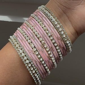 May include: A collection of pink bangles with intricate detailing. The bangles are adorned with small, sparkling embellishments and are stacked on a wrist. The overall aesthetic is elegant and decorative, suitable for special occasions.