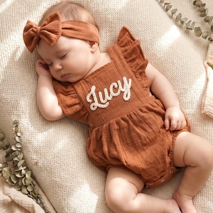 May include: A sleeping baby wearing a rust-colored romper with the name "Lucy" embroidered on the front. The baby also wears a matching headband with a bow. The romper has ruffled sleeves.