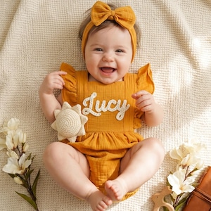 Könnte beinhalten: Ein lächelndes Baby in einem senfgelben Strampler mit dem Namen "Lucy" auf der Vorderseite. Das Outfit beinhaltet ein passendes Stirnband und wird mit einem gehäkelten Sonnen-Spielzeug ergänzt. Das Baby liegt auf einer cremefarbenen Decke.