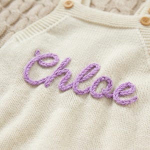 May include: Cream-colored knitted baby romper with the name "Chloe" embroidered in lavender thread. The romper features wooden buttons and a cable knit design on the straps. The soft, textured fabric suggests a cozy and comfortable garment.