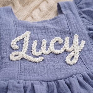 May include: Close-up of a light blue dress with the name "Lucy" embroidered in white, cursive letters. The dress has a ruffled neckline and a textured fabric. The background is a soft, neutral color.