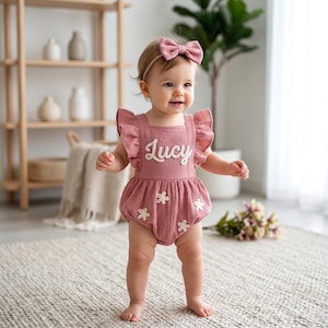 May include: A dusty rose baby romper with white floral accents and the name "Lucy" embroidered on the front. The romper has ruffled sleeves and is paired with a matching bow headband. The baby is standing on a neutral-colored rug.