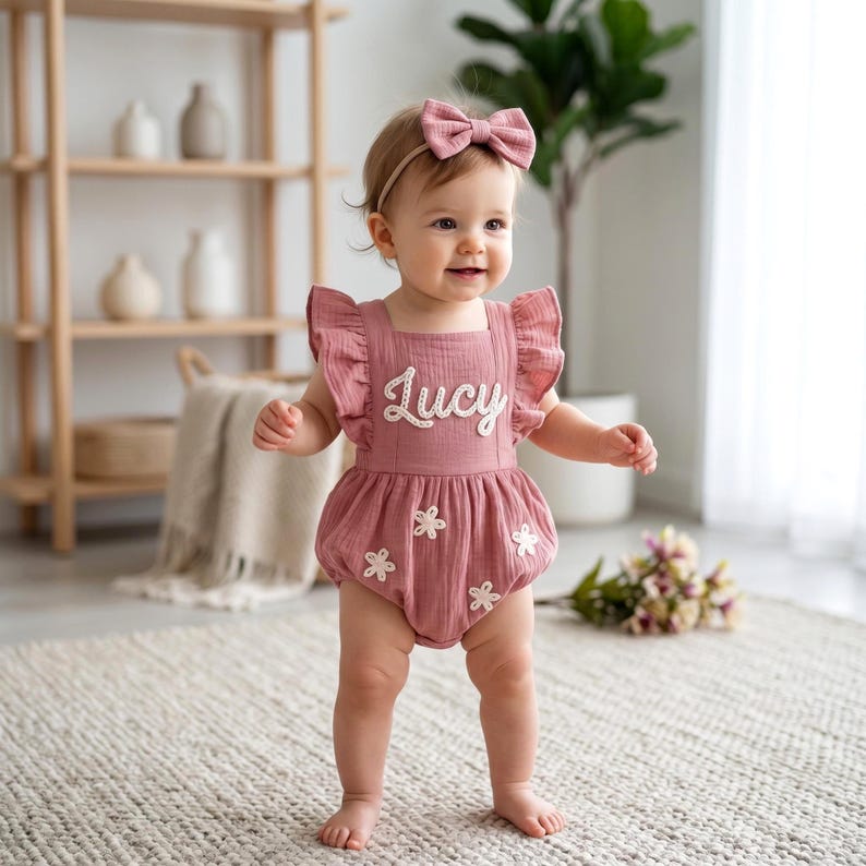 May include: A dusty rose baby romper with white floral accents and the name "Lucy" embroidered on the front. The romper has ruffled sleeves and is paired with a matching bow headband. The baby is standing on a neutral-colored rug.