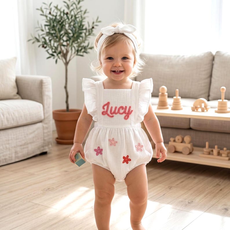 May include: A white baby romper with the name "Lucy" embroidered in pink script. The romper features ruffled sleeves and is decorated with pink and red flower appliqu&eacute;s. A matching white headband completes the outfit.