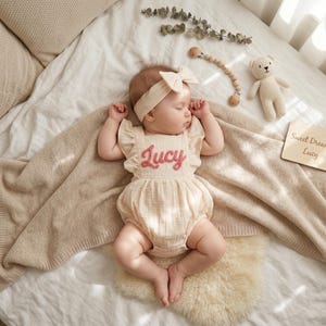 May include: A sleeping baby wearing a beige romper with the name "Lucy" embroidered in pink. The baby is also wearing a matching headband with a bow. A wooden sign that says "Sweet Dreams Lucy" is next to the baby.