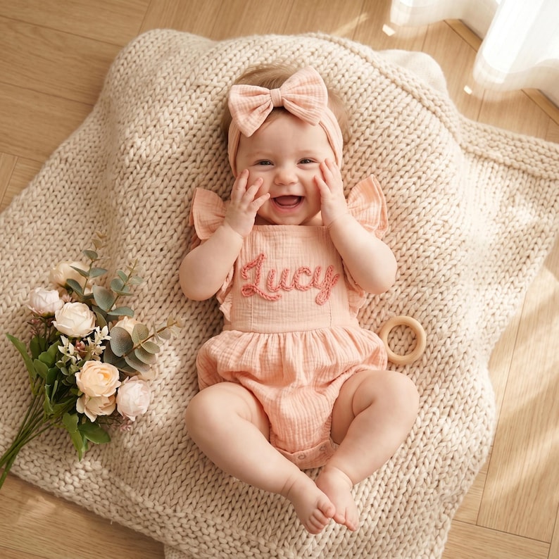 May include: A baby wearing a peach-colored romper with the name "Lucy" embroidered on the front, paired with a matching bow headband. The baby is lying on a cream-colored knitted blanket, with a bouquet of flowers and a wooden toy nearby.