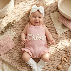 May include: A smiling baby wearing a pink knitted romper with the name "Chloe" in white script. The romper has wooden button details and ruffle trim. The baby is wearing a white bow headband and socks, lying on a textured blanket.