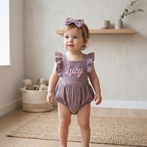 May include: A baby wearing a mauve romper with ruffled straps and the name "Lucy" embroidered on the front. The baby also wears a matching headband with a bow. The background includes a woven basket, a wooden shelf, and a neutral-toned wall.
