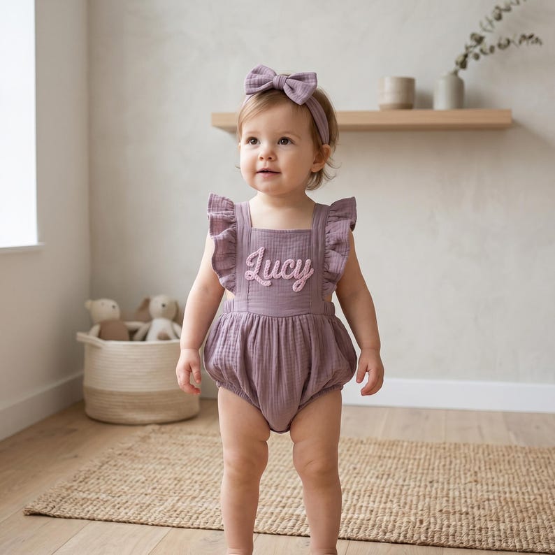 May include: A baby wearing a mauve romper with ruffled straps and the name "Lucy" embroidered on the front. The baby also wears a matching headband with a bow. The background includes a woven basket, a wooden shelf, and a neutral-toned wall.