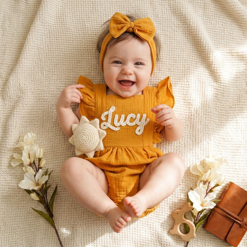 May include: A smiling baby wearing a mustard yellow romper with the name "Lucy" embroidered on the front. The outfit includes a matching headband with a bow. A crocheted sun toy and flowers are nearby.