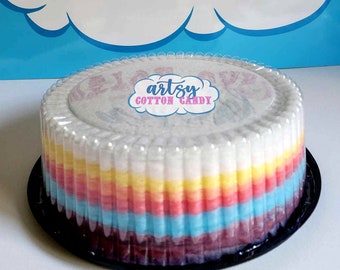 Könnte beinhalten: Ein regenbogenfarbener, mehrschichtiger Baumwollzuckerkuchen in einem durchsichtigen Plastikbehälter mit schwarzem Boden. Die Oberseite des Kuchens ist mit einem weißen Papierkreis mit dem Text "artsy cotton candy" verziert.