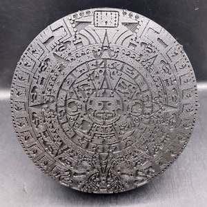 Puede incluir: Una escultura circular en relieve negro con intrincados diseños y símbolos. La escultura recuerda a la piedra del calendario azteca.