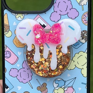 May include: A white and pink phone case with a glitter phone grip featuring a Mickey Mouse head with a pink bow and white icing dripping down. The case has a blue background with a pattern of Mickey Mouse heads, ice cream cones, donuts, and other Disney-themed icons.