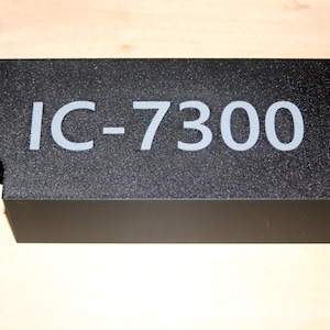 May include: A rectangular black cover with a textured finish featuring the white text IC-7300 printed on the top surface. The cover includes a small circular cutout on the left side, designed to accommodate a cable or connector for radio equipment.